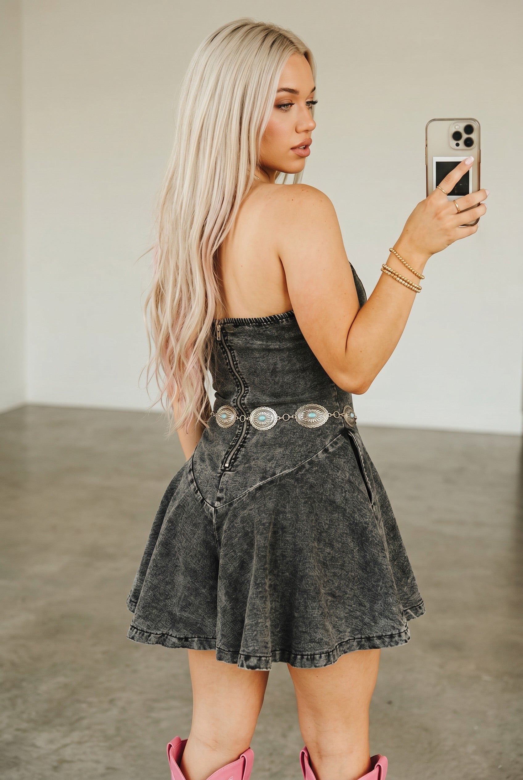 Woman taking a mirror selfie wearing a black denim dress with silver belt buckles.