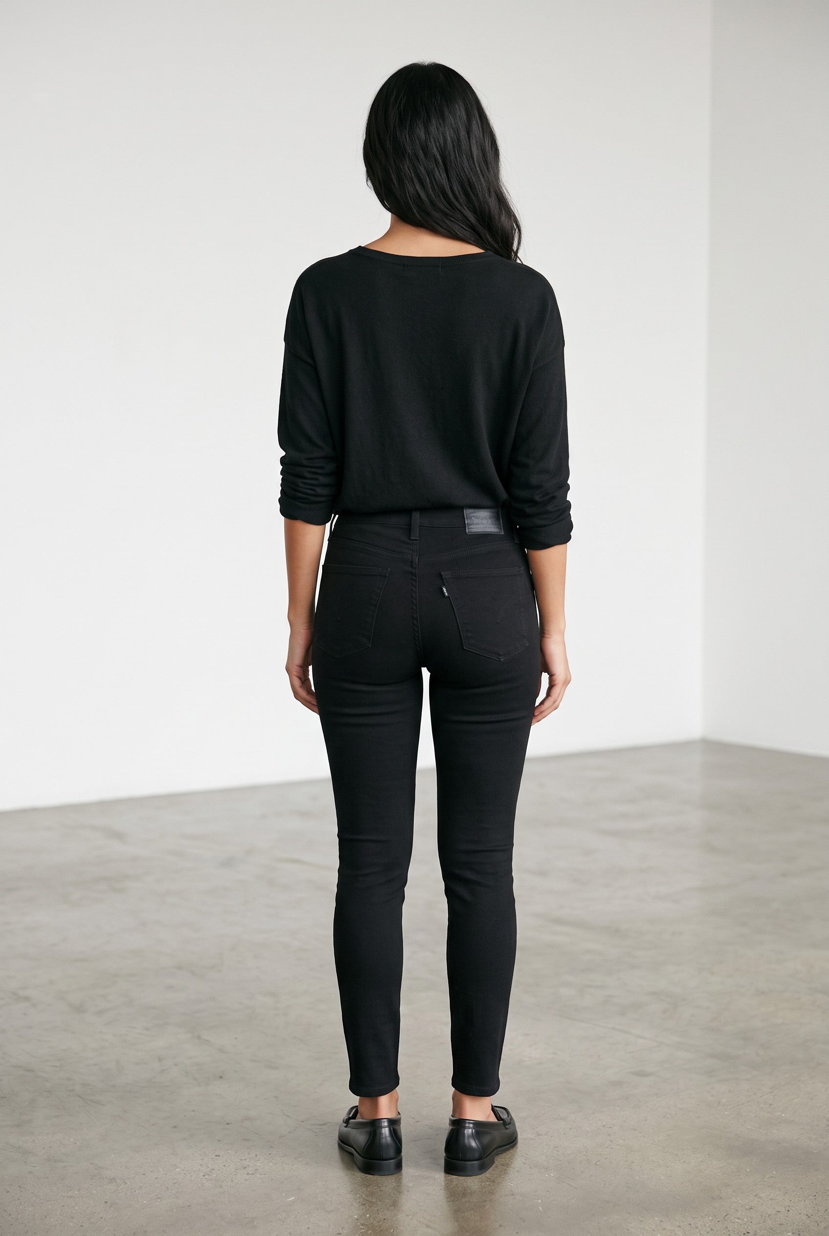 A woman viewed from behind wearing a black long-sleeve top and black skinny jeans, standing in a minimalist studio with a white wall and gray concrete floor.