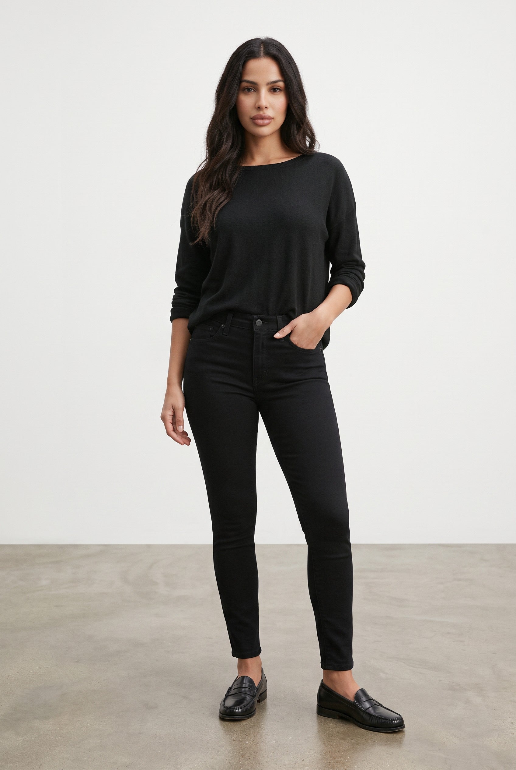 A woman modeling a black long-sleeve top and black skinny jeans, paired with black loafers, standing against a plain light gray studio background.