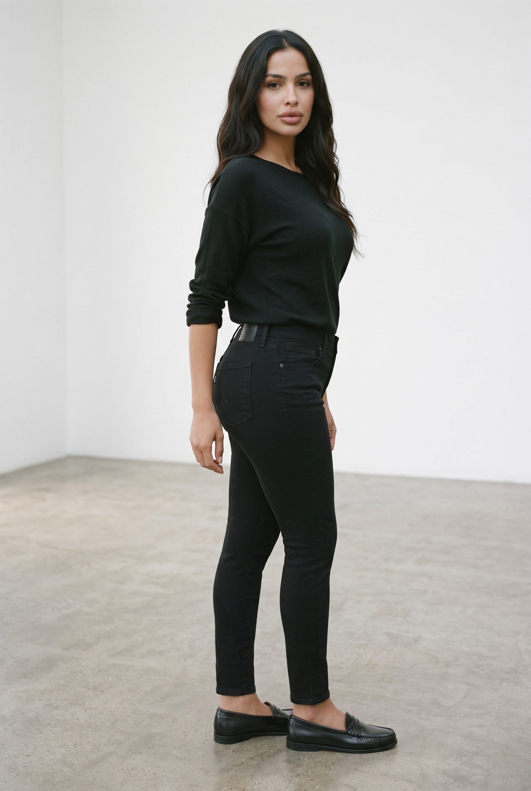 A woman modeling a black long-sleeve top and black skinny jeans, standing in a minimalist studio with a white wall and gray concrete floor.