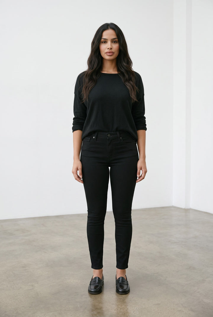 A woman stands in a minimalist studio wearing a black long-sleeve top, black skinny jeans, and black leather loafers.